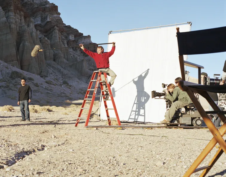 A film crew on set with a boom microphone and a white backdrop in a desert.